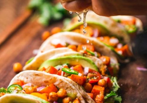 Several tacos with chickpeas, avocado, cilantro, and more are on a wooden board. A hand squeezes lime juice over them.