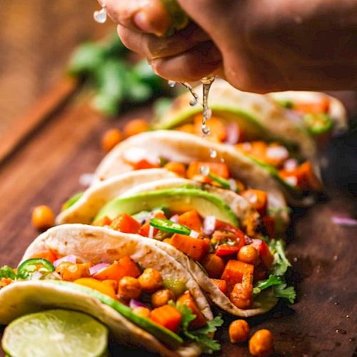 Several tacos with chickpeas, avocado, cilantro, and more are on a wooden board. A hand squeezes lime juice over them.