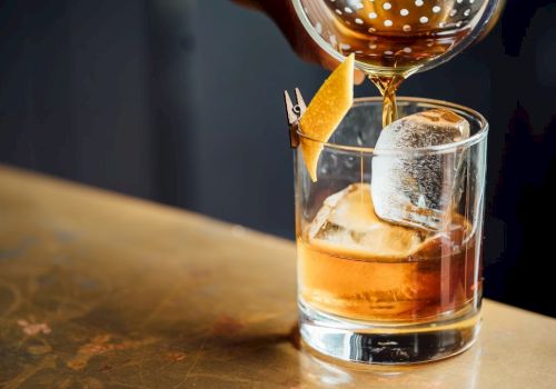 A cocktail being poured into a glass with ice and a twist of citrus peel garnish on the rim, set on a countertop.