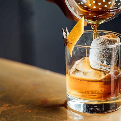 A cocktail being poured into a glass with ice and a twist of citrus peel garnish on the rim, set on a countertop.