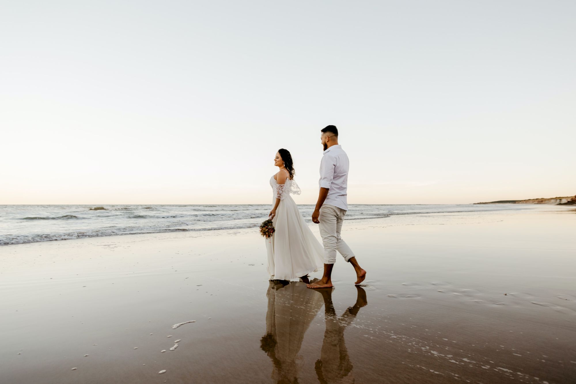 A couple walks barefoot on a beach with soft waves and a pastel sky, reflecting their silhouettes on the wet sand.