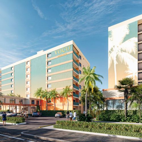 A modern hotel with colorful exterior and palm trees, surrounded by a street and parked cars on a sunny day.