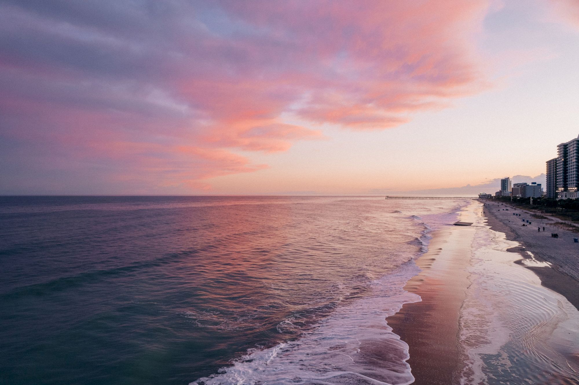 A coastal scene at sunset shows pink clouds, calm waves, and a sandy beach with buildings lining the shore in the background.