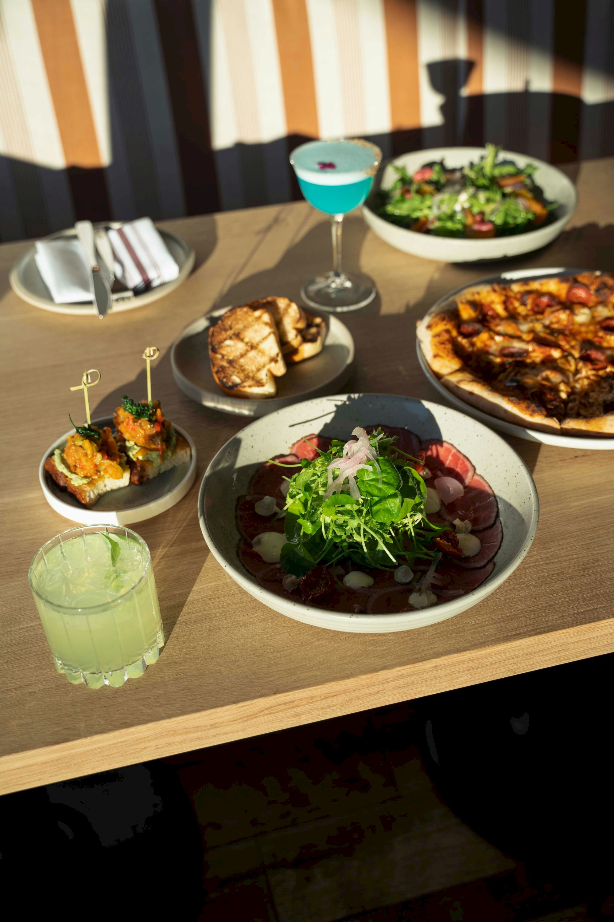 A table with various dishes and drinks: salads, pizza, appetizers, a blue cocktail, and a green beverage, set for a meal.