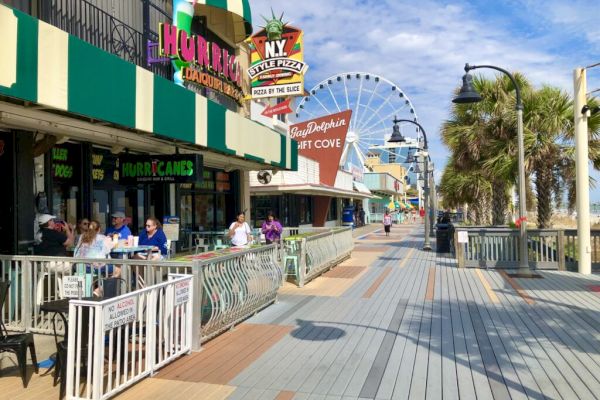 A lively boardwalk scene with people dining, colorful storefronts, and a Ferris wheel in the background under a partly cloudy sky.