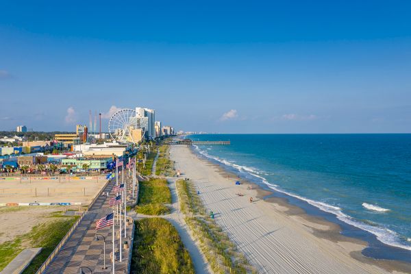 A beach scene with a boardwalk, flags, hotels, a ferris wheel, and a sandy shoreline meeting the ocean under a clear blue sky.