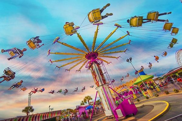 The image shows a colorful amusement park with people enjoying a spinning swing ride under a vibrant sky.