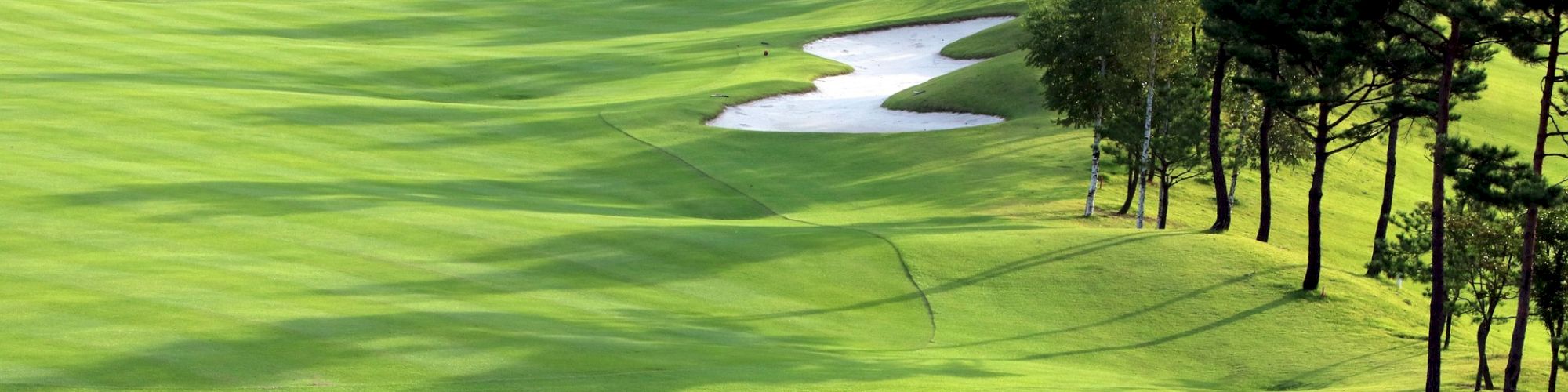 The image shows a scenic golf course fairway with lush green grass, sand bunkers, and trees lining the edges under clear skies.