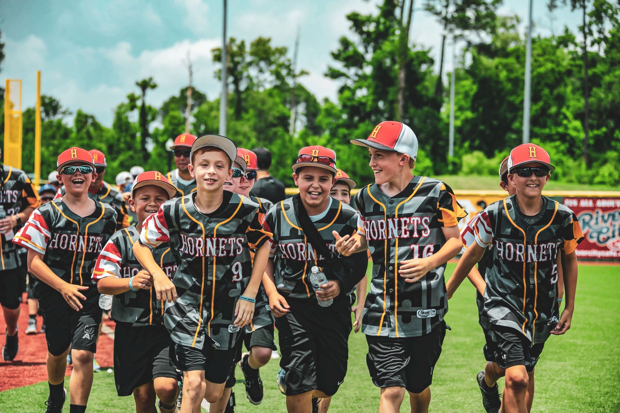 A group of young baseball players in matching uniforms and caps are running on a field, smiling and appearing excited.