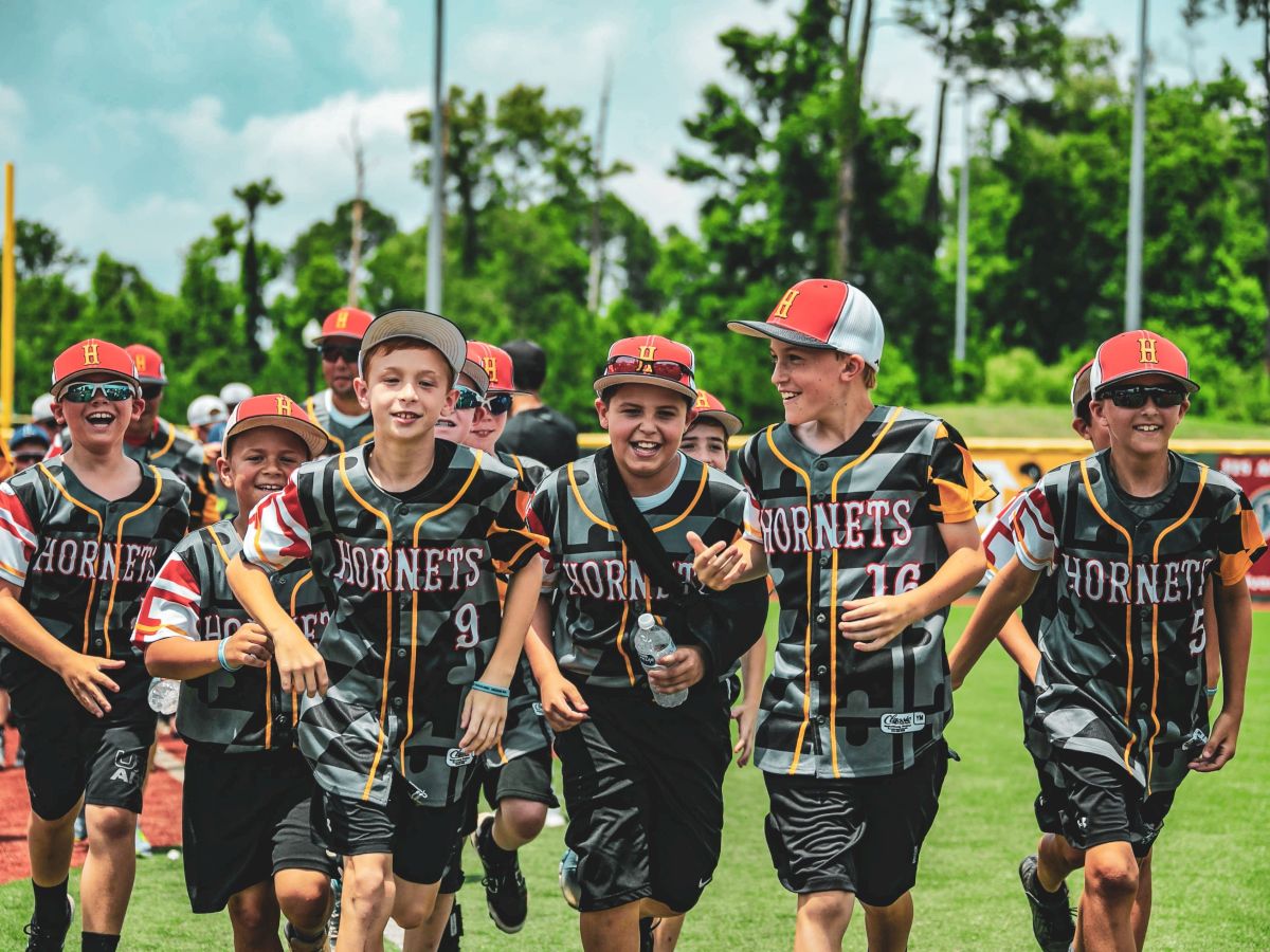 A group of young baseball players in "Hornets" uniforms run together on a field, smiling and enjoying their time outdoors.