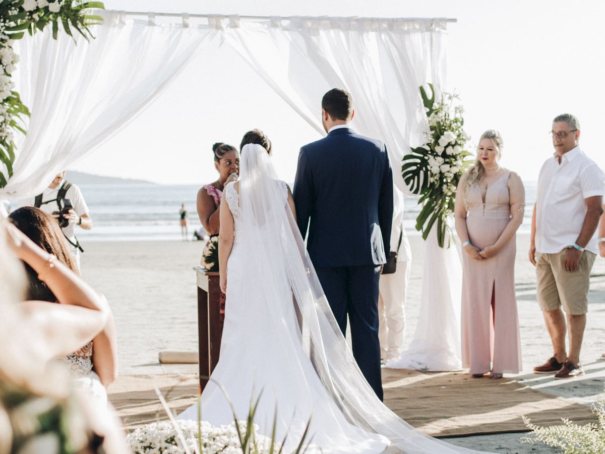 A beach wedding ceremony with a couple standing under a white canopy, surrounded by attendants and guests, with the ocean in the background.