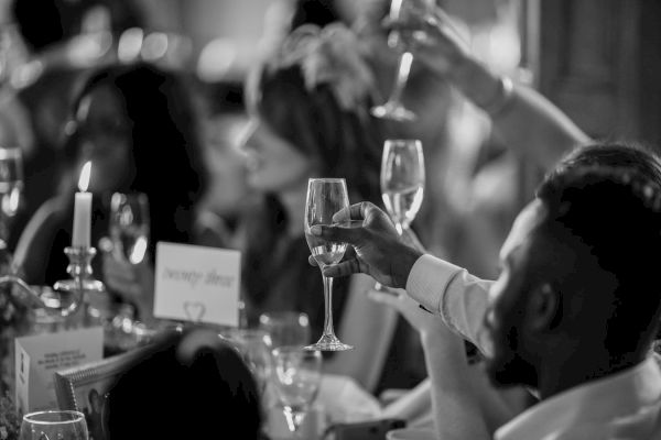 People are raising glasses in a celebratory toast at an indoor event. The scene is in black and white, creating an elegant atmosphere.