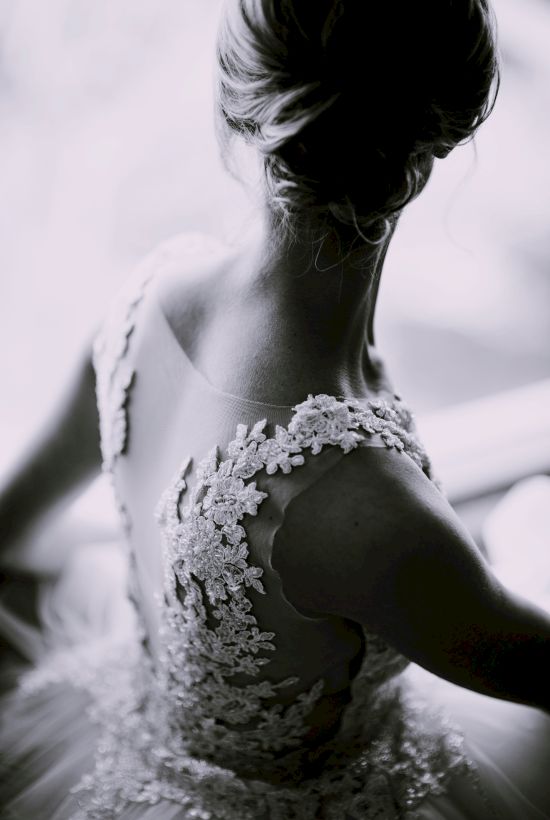 A black and white image of a woman in an intricately detailed dress, viewed from the back, with a blurred background.