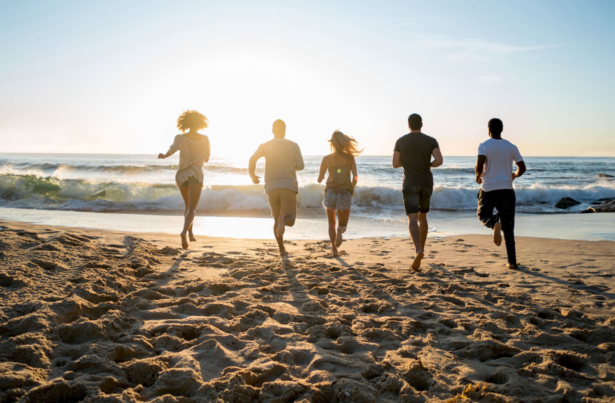 A group of five people is running toward the ocean on a sandy beach, with the sun setting in the background.