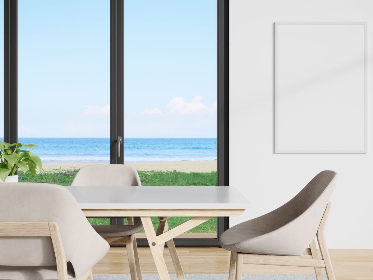 A modern dining area with neutral chairs and a table overlooks a beach scene through large windows. A blank frame hangs on the wall.