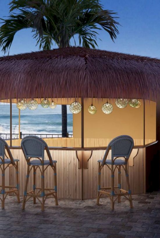 Tiki bar with straw roof and bar stools, by the ocean under palm trees, surrounded by colorful striped umbrellas, at sunset.