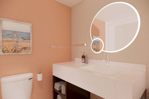 The image shows a bathroom with a round, illuminated mirror, a sink, a toilet, and a wall art piece next to a coral-colored wall.