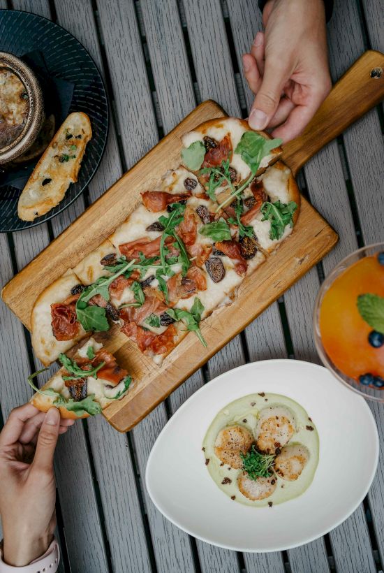 A flatbread with arugula and tomatoes on a wooden board, a bowl of soup, and a orange drink with blueberries are on the table.