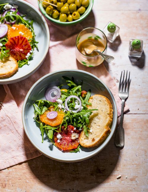 A salad with arugula, orange slices, onions, bread, and dressing. Green olives in a separate bowl and a fork on the side complete the setting.