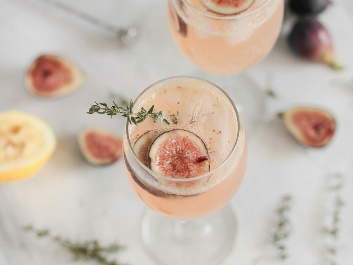 Two glasses of a pink drink with fig slices and thyme sprigs, surrounded by fig pieces and a lemon slice on a marble surface.