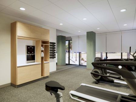 The image shows a gym with exercise machines, wall cabinets, towels, and windows with blinds in a well-lit room.