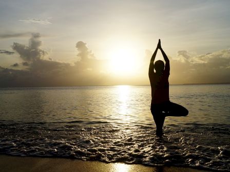 A person practices yoga on the beach at sunset, striking a tree pose against a serene ocean backdrop.