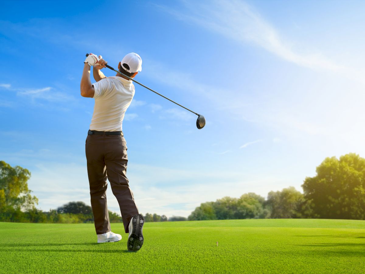 A golfer swings a club on a lush green course under a blue sky. Trees line the background, and the sun is shining brightly.