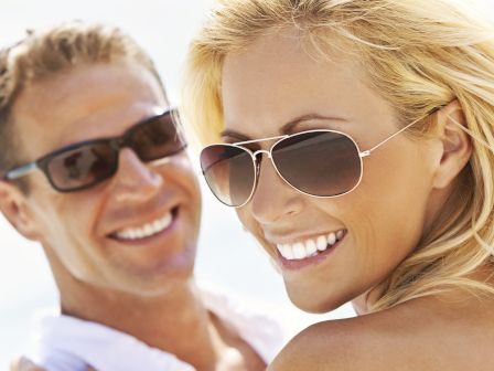 A smiling blonde woman and a man, both wearing sunglasses, enjoying a sunny day outdoors, with a bright background.