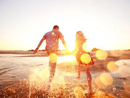 A couple holding hands runs through shallow water at sunset, creating splashes and lens flares in a warm, glowing atmosphere.